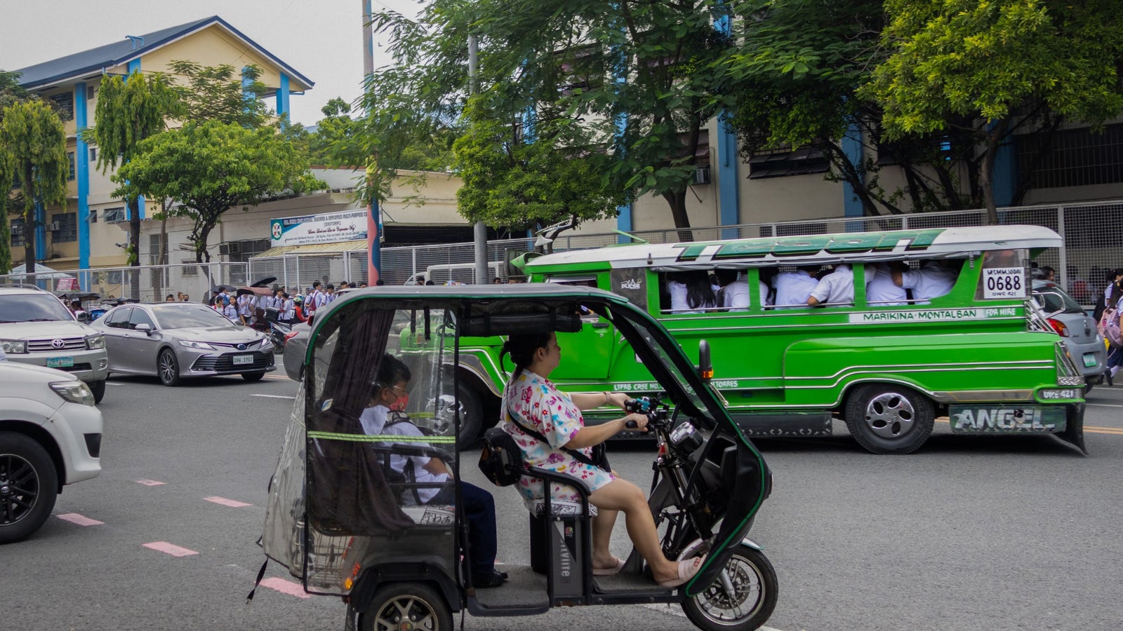 A photo of a woman driving an e-tricycle across a busy road.